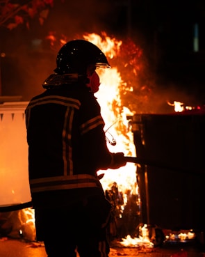 A firefighter in protective gear stands in front of a large, outdoor fire, holding a hose. The flames illuminate the scene, creating an intense and urgent atmosphere.