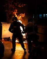 Two firefighters wearing protective gear are actively managing a blaze in an outdoor setting at night. The fire is emanating bright flames from a burning object, possibly a trash bin, casting a warm glow and illuminating the firefighters. The surrounding area is dark, with some trees visible.