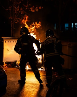 Two firefighters wearing protective gear are actively managing a blaze in an outdoor setting at night. The fire is emanating bright flames from a burning object, possibly a trash bin, casting a warm glow and illuminating the firefighters. The surrounding area is dark, with some trees visible.