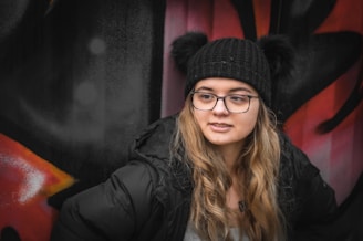 A young person wearing a black knitted hat with pompoms is leaning against a wall with colorful graffiti art. They have long, wavy blonde hair, glasses, and are dressed in a black jacket. The background graffiti features abstract shapes in shades of red, black, and white.