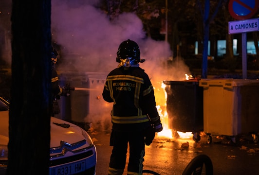 Firefighters stand in front of a burning trash bin on a street, wearing protective gear with reflective stripes. The scene is illuminated by the flames and there is smoke rising into the night sky. A parked car and road signs are visible nearby.