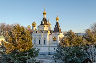 An ornate building with golden domes and intricate architectural details is surrounded by evergreen trees. The structure exhibits traditional Eastern European design elements and is basking in sunlight under a clear blue sky.