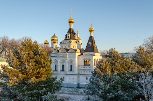 An ornate building with golden domes and intricate architectural details is surrounded by evergreen trees. The structure exhibits traditional Eastern European design elements and is basking in sunlight under a clear blue sky.