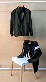 Two jackets are displayed against a plain background. One jacket is hanging on a wooden hanger from a rod while the other is draped over the back of a chair with light wooden legs and white seat. Both jackets appear to have a smooth texture and a similar design with a front zipper closure.