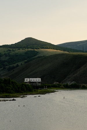 A serene landscape featuring a small body of water in the foreground bordered by thick greenery. In the distance, gently rolling hills are bathed in soft light, with a small, unassuming structure positioned on the edge of the water, partially surrounded by vegetation.