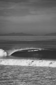 A black and white photograph capturing a powerful ocean wave curling as it approaches the shore. A lone surfer is seen paddling in the wave, set against a backdrop of distant, hazy mountains under a cloudy sky.