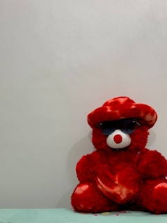 A red teddy bear with a white snout and eyes, wearing dark sunglasses and a matching red hat, sits against a plain light-colored wall. The bear is positioned on a surface that appears to be a table.