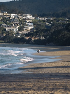 A lone surfer carrying a surfboard walks along a sandy beach. The waves gently crash against the shore, and in the background, a hill is covered with numerous modern houses nestled among trees.