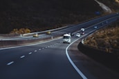 A highway stretches into the distance with multiple lanes. Several vehicles, including cars and a bus, are traveling in both directions. The surroundings appear to be hilly with greenery on one side and open space on the other.