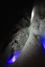 A person equipped with climbing gear, including a helmet and crampons, ascends an icy vertical surface in a dark environment. The ice has a mix of smooth and jagged textures, illuminated by a soft white light while purple and blue lights add an ethereal glow from below. The climber is progressing carefully in a challenging and frozen setting.