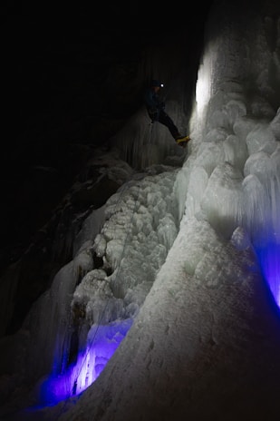 A person equipped with climbing gear, including a helmet and crampons, ascends an icy vertical surface in a dark environment. The ice has a mix of smooth and jagged textures, illuminated by a soft white light while purple and blue lights add an ethereal glow from below. The climber is progressing carefully in a challenging and frozen setting.