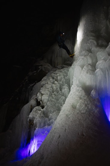 A person equipped with climbing gear, including a helmet and crampons, ascends an icy vertical surface in a dark environment. The ice has a mix of smooth and jagged textures, illuminated by a soft white light while purple and blue lights add an ethereal glow from below. The climber is progressing carefully in a challenging and frozen setting.