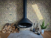 A modern, black hanging fireplace against a light brick wall, with a neatly stacked pile of firewood to the left. To the right, fireplace tools, a potted small green plant in a white vase, and a decorative cylindrical log with branches are arranged on a tiled floor.