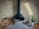 A modern, black hanging fireplace against a light brick wall, with a neatly stacked pile of firewood to the left. To the right, fireplace tools, a potted small green plant in a white vase, and a decorative cylindrical log with branches are arranged on a tiled floor.