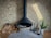 A modern, black hanging fireplace against a light brick wall, with a neatly stacked pile of firewood to the left. To the right, fireplace tools, a potted small green plant in a white vase, and a decorative cylindrical log with branches are arranged on a tiled floor.