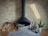 A modern, black hanging fireplace against a light brick wall, with a neatly stacked pile of firewood to the left. To the right, fireplace tools, a potted small green plant in a white vase, and a decorative cylindrical log with branches are arranged on a tiled floor.