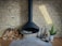 A modern, black hanging fireplace against a light brick wall, with a neatly stacked pile of firewood to the left. To the right, fireplace tools, a potted small green plant in a white vase, and a decorative cylindrical log with branches are arranged on a tiled floor.