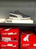A black and white Puma sneaker is displayed on a backlit shelf. Below the shoe, several red Puma shoeboxes are stacked. The boxes are labeled with 'SOFTFOAM+' and the Puma logo.