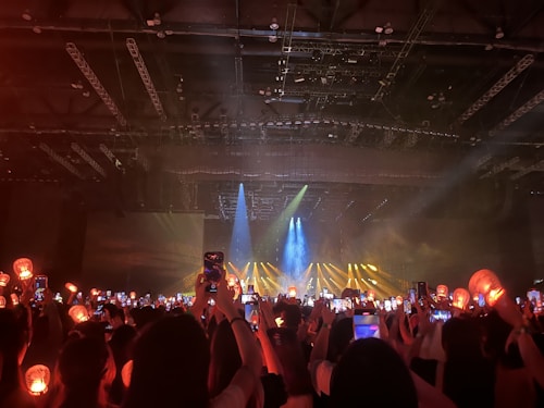 A dimly lit indoor concert venue filled with an audience holding up lit devices and lantern-like objects, capturing a performance on stage. Bright spotlights and blue and yellow beams illuminate the stage area, creating a lively atmosphere.