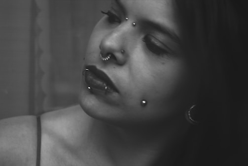 A black and white portrait of a person with multiple facial piercings, including a nose ring, lip ring, and cheek studs. The individual has a contemplative expression and dark lipstick. Their head is slightly tilted to the side, and they have long hair falling to one side.