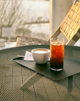 A table with a textured metal surface holds a tray with a cup of cappuccino and a tall glass of iced coffee. The background suggests an outdoor setting with sunlight filtering through, creating a warm and relaxed ambiance.
