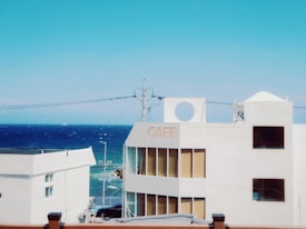 A white building with a sign reading 'CAFE' overlooks a vibrant blue ocean under a clear sky. Several windows reflect the sunlight, and there are power lines crossing the view. The scene captures a serene coastal ambiance.
