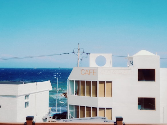 A white building with a sign reading 'CAFE' overlooks a vibrant blue ocean under a clear sky. Several windows reflect the sunlight, and there are power lines crossing the view. The scene captures a serene coastal ambiance.