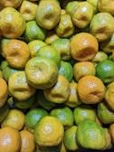 A close-up view of a pile of citrus fruits, predominantly featuring limes and small oranges. The fruits exhibit a mix of green and yellowish colors, with some showing minor surface blemishes.