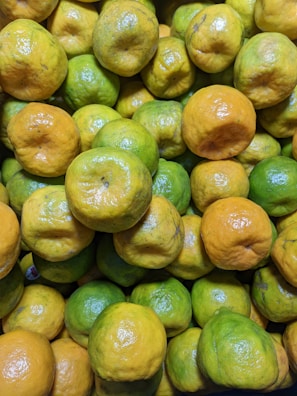 A close-up view of a pile of citrus fruits, predominantly featuring limes and small oranges. The fruits exhibit a mix of green and yellowish colors, with some showing minor surface blemishes.