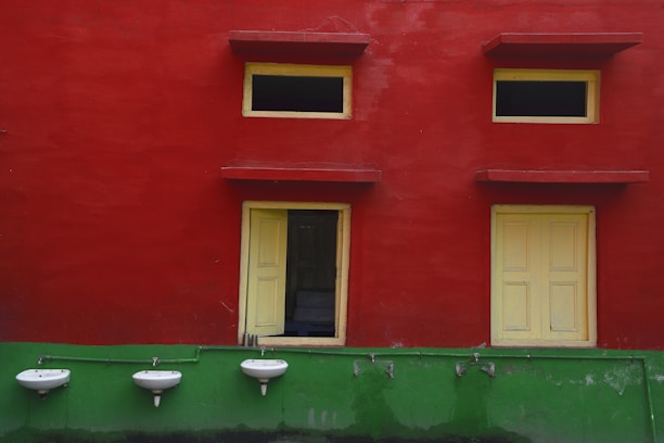 A brightly painted wall with two small windows and a door, all with yellow frames, against a red backdrop. Below the windows, there are three white, wall-mounted sinks attached to a green-painted lower section of the wall. The image features a stark contrast between the vibrant red and green colors.