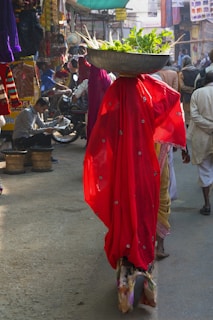 A person wearing a vibrant red sari carries a large metal bowl filled with leafy greens and vegetables on their head while walking through a bustling market street. The street is lively with people and various shops displaying colorful textiles and goods. Another person reads a newspaper, seated near a parked motorcycle.