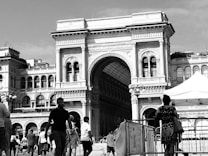 A historic arch structure dominates the scene, with intricate architectural details and a vaulted roof extending into the background. People are walking in front of the building, suggesting a busy public area. The scene appears to be monochrome, adding a timeless quality.