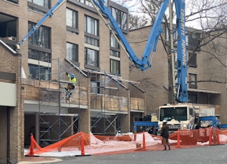 A construction site in front of a multi-story brick building, featuring scaffolding and a construction worker wearing a yellow safety vest and helmet. A large blue concrete pumping truck is parked nearby, extending its arm toward the building. Orange safety netting and cones cordon off the area.