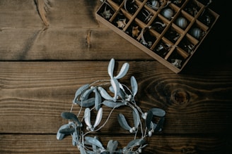 A wooden table surface with a decorative wreath made of grey leaves and berries positioned in the lower part of the image. In the upper right corner, a wooden box with compartments is filled with small decorative items such as acorns and twigs.
