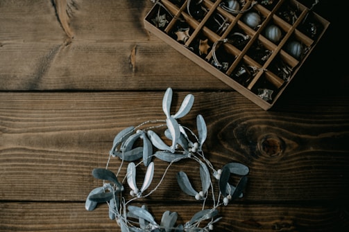 A wooden table surface with a decorative wreath made of grey leaves and berries positioned in the lower part of the image. In the upper right corner, a wooden box with compartments is filled with small decorative items such as acorns and twigs.