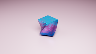 A uniquely shaped geometric object stands on a plain, light-colored surface. The object features a twisted form and is predominantly blue with purple and pink hues. It has a reflective, smooth texture, creating an abstract and artistic appearance.