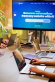 A conference room setting with several laptops on a large table, each being used by a person. A large screen displays a blue interface with the text 'Generate ad creatives from any website with AI'. A stainless steel water bottle and a conference phone are also visible on the table.