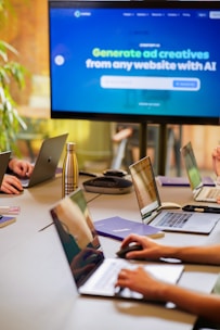 A conference room setting with several laptops on a large table, each being used by a person. A large screen displays a blue interface with the text 'Generate ad creatives from any website with AI'. A stainless steel water bottle and a conference phone are also visible on the table.