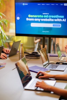 A conference room setting with several laptops on a large table, each being used by a person. A large screen displays a blue interface with the text 'Generate ad creatives from any website with AI'. A stainless steel water bottle and a conference phone are also visible on the table.