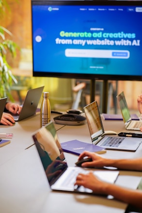 A conference room setting with several laptops on a large table, each being used by a person. A large screen displays a blue interface with the text 'Generate ad creatives from any website with AI'. A stainless steel water bottle and a conference phone are also visible on the table.