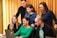 Six people are gathered around a laptop in an office setting. They appear focused and engaged, possibly collaborating on a project. The group includes both men and women, and some are seated while others stand. A yellow curtain is in the background, adding a warm tone to the scene.