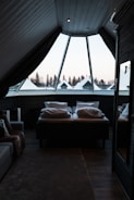 A cozy attic bedroom featuring a large triangular window that overlooks a snowy landscape with pine trees. The room includes a comfortable bed covered with pillows and a thick blanket, and a soft, textured carpet on the wooden floor. Dark curtains partially cover the window, creating a warm and secluded atmosphere.