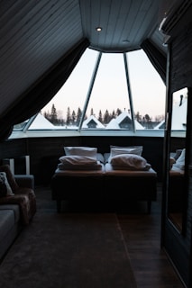 A cozy attic bedroom featuring a large triangular window that overlooks a snowy landscape with pine trees. The room includes a comfortable bed covered with pillows and a thick blanket, and a soft, textured carpet on the wooden floor. Dark curtains partially cover the window, creating a warm and secluded atmosphere.
