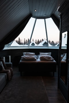 A cozy attic bedroom featuring a large triangular window that overlooks a snowy landscape with pine trees. The room includes a comfortable bed covered with pillows and a thick blanket, and a soft, textured carpet on the wooden floor. Dark curtains partially cover the window, creating a warm and secluded atmosphere.
