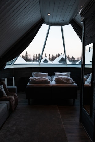 A cozy attic bedroom featuring a large triangular window that overlooks a snowy landscape with pine trees. The room includes a comfortable bed covered with pillows and a thick blanket, and a soft, textured carpet on the wooden floor. Dark curtains partially cover the window, creating a warm and secluded atmosphere.