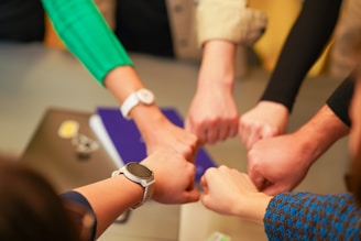Several people form a circle, joining their fists together in a gesture of unity and teamwork. Various wristwatches and clothing styles are visible, suggesting a diverse group.
