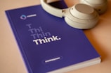 A pair of light-colored headphones rests on top of a blue notebook with the word 'Think' prominently displayed on the cover. The background is a neutral color, providing a calm atmosphere.