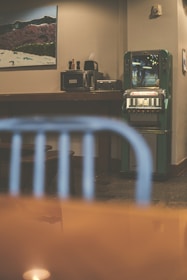A retro-styled indoor setting featuring a green vintage vending machine against a beige wall. On the left, there is a wooden counter with a few appliances, including a coffee maker and a paper towel holder. Above the counter hangs a framed picture depicting a mountainous landscape. In the foreground, there's a blurred blue metal chair, and the reflection of a light is visible on a glossy surface.