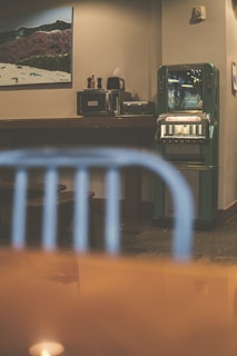 A retro-styled indoor setting featuring a green vintage vending machine against a beige wall. On the left, there is a wooden counter with a few appliances, including a coffee maker and a paper towel holder. Above the counter hangs a framed picture depicting a mountainous landscape. In the foreground, there's a blurred blue metal chair, and the reflection of a light is visible on a glossy surface.