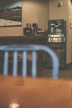 A retro-styled indoor setting featuring a green vintage vending machine against a beige wall. On the left, there is a wooden counter with a few appliances, including a coffee maker and a paper towel holder. Above the counter hangs a framed picture depicting a mountainous landscape. In the foreground, there's a blurred blue metal chair, and the reflection of a light is visible on a glossy surface.